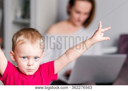 A Young Mother Sits On A Couch And Types On A Laptop While Her Little Son Indulges And Poses For The