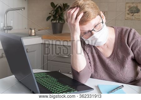 A Tired Middle-aged Woman In A Medical Mask And Glasses Sits At A Table With A Laptop In A Cozy Kitc