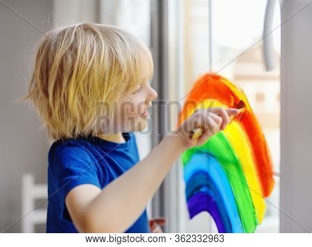 Little Boy Sitting At Window With Drawing Rainbow While Coronavirus Quarantine. Rainbow Sign Is Symb