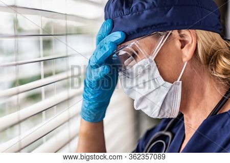 Stressed Female Doctor or Nurse On Break At Window Wearing Medical Face Mask and Goggles.