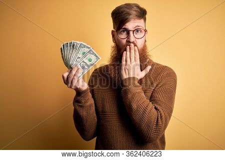 Redhead Irish man with beard holding a bunch of 1 dolla banknotes over yellow background cover mouth with hand shocked with shame for mistake, expression of fear, scared in silence, secret concept