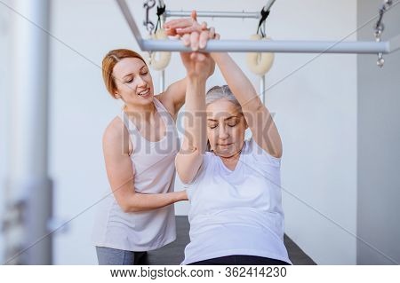 Older Woman Doing Exercise On Pilates Chair Equipment. Young Female Personal Trainer Helping Senior 