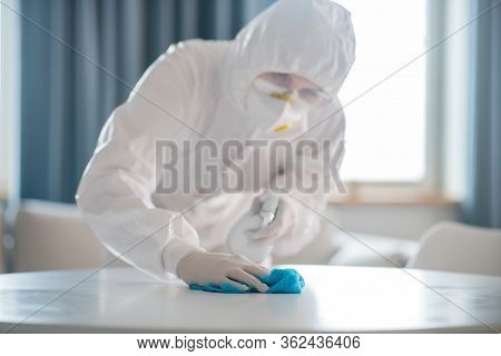 Woman In White Workwear And Protective Gloves Looking Involved While Disinfecting The Table Surface