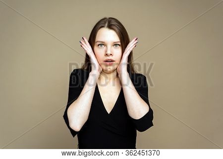 A Shoked Girl Is Shruging. A Girl Is Open-eyes On The Brown Background. A Girl With Hands In The Fac