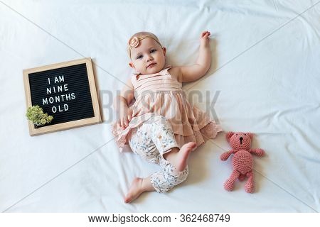 9 Nine Months Old Baby Girl Laying Down On White Background With Letter Board And Teddy Bear. Flat L