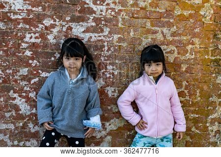Asian Identical Female Twins Child Wearing Jacket During Cold Weather And Standing On An Old Brick W