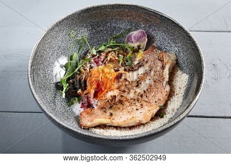 Sea bass fillet with shiitake mushrooms in gray bowl top view. Grouper meat with vegetables, greenery and violet flowers decoration. Delicious porgy, seafood with veggies, served restaurant meal