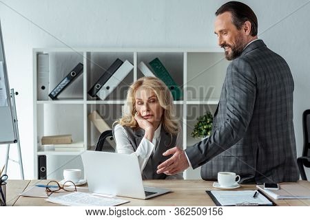 Smiling Businessman Pointing With Hand At Laptop Near Businesswoman At Table