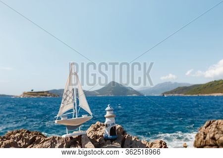 A Wooden Sailing Ship With A Lighthouse Stand On A Rocky Shore Against The Sea. Against The Backdrop
