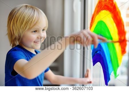 Little Boy Sitting At Window With Drawing Rainbow While Coronavirus Quarantine. Rainbow Sign Is Symb