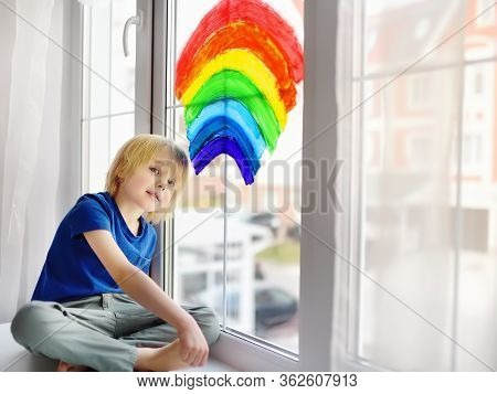 Little Boy Sitting At Window With Drawing Rainbow While Coronavirus Quarantine. Rainbow Sign Is Symb