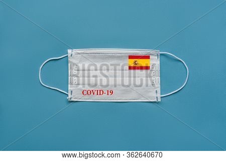 Medical Mask With The Flag Of Spain And The Inscription Covid-19 On A Blue Background