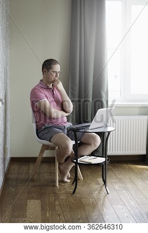 The Man In The Pink Polo Siiting By The Small Table And Working At Home On Laptop. Man In Quarantine
