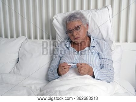 Senior Woman Sittnig In Bed Checking Her Temperature On Thermometer. Modern Bedroom In White Colors.