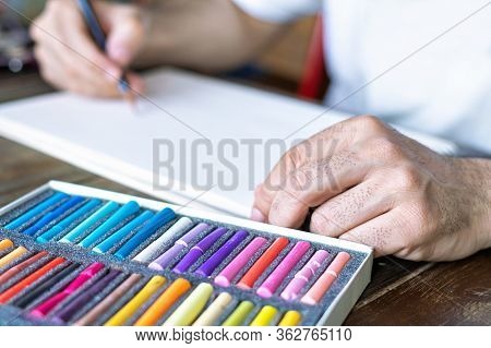 Artist Painting With A Pastel Crayon Chalk On A White Sheet Of Paper And Box With Pastel Chalks In D
