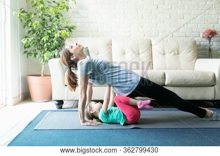 Profile View Of A Little Girl Getting Behind Her Mother While She Practices Yoga And Holds A Reverse