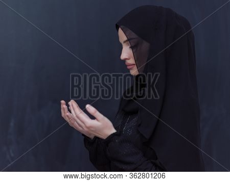 portrait of beautiful muslim woman in fashionable dress with hijab making traditional prayer to God, keeps hands in praying gesture isolated on black chalkboard background representing modern islam 