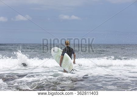 The Surf Enters The Water. Male Surfer Entering The Sea With His Board In A Black Surfing Suit. Tene