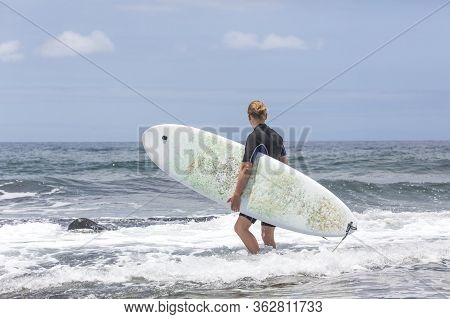 The Surf Enters The Water. Male Surfer Entering The Sea With His Board In A Black Surfing Suit. Tene
