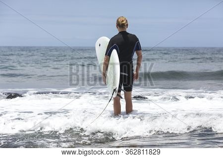 The Surf Enters The Water. Male Surfer Entering The Sea With His Board In A Black Surfing Suit. Tene