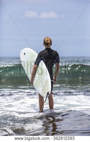 The Surf Enters The Water. Male Surfer Entering The Sea With His Board In A Black Surfing Suit. Tene