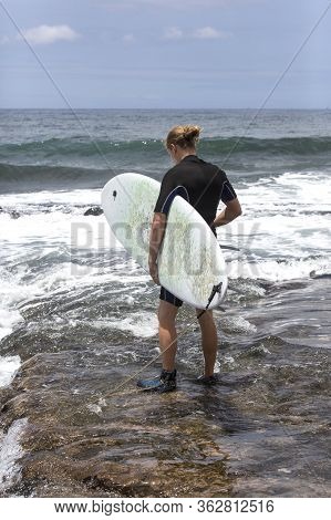 The Surf Enters The Water. Male Surfer Entering The Sea With His Board In A Black Surfing Suit. Tene