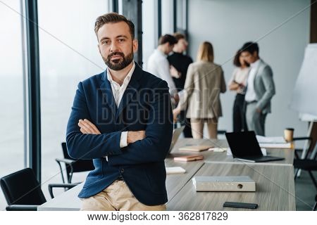 Attractive young confident businessman in formal wear sittng in the office with his colleagues on a background, arms folded