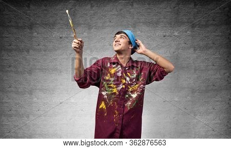 Smiling Young Painter Artist Pointing Upwards With Paintbrush In Hand. Portrait Of Happy Painter On 