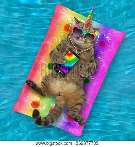 The Beige Cat Unicorn In Headphones And Heart Shaped Sunglasses With A Smartphone Is Lying On A Mult