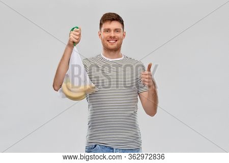 food, sustainability and eco living concept - smiling young man in striped t-shirt holding reusable string bag with bananas and showing thumbs up over grey background