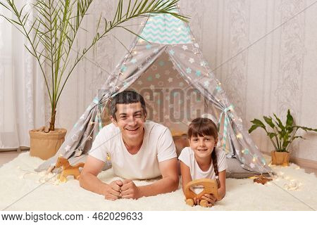 Indoor Shot Of Smiling Handsome Caucasian Man Wearing White T Shirt Lying On Floor With His Little D