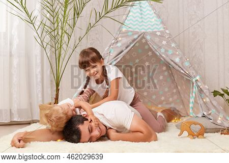 Indoor Shot Of Happy Smiling Man Wearing White T-shirt Posing With His Children On Floor Near Wigwam