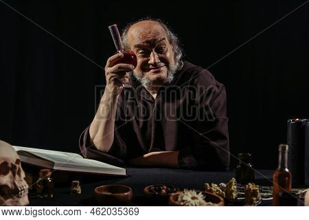 Cheerful Alchemist Showing Flask With Potion Near Magic Cookbook Isolated On Black.