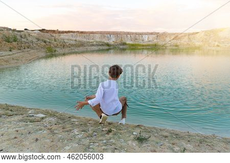 A Boy Of 8-9 Years Old Throws Stones Into The Water From The Shore, Fun In Nature.