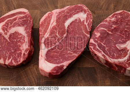 Close Up Three Raw Marbled Ribeye Beef Steaks Over End Grain Cutting Board Of Wooden Butcher Block, 