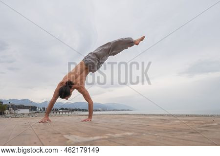 Shirtless Caucasian Man Doing Backflip On Pebble Beach.