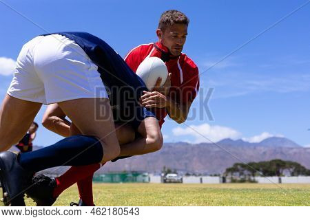 Low angle side view of two Caucasian male rugby players from opposing teams wearing team uniforms, in a tackle on a rugby pitch during a match, with blue sky in the background