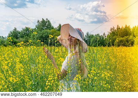 Beautiful Girl In Wide-brimmed Hat In A Flowering Field With White Dress. Sunbeams, Glare, Light Lea