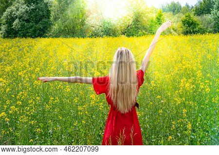 Beautiful Girl In A Flowering Field In A Red Dress With Arms Wide Open. Sunbeams, Glare, Light Leaks