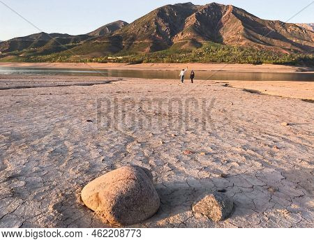 Beautiful Landscape With Setting Sun: Mountains, Lake, Cracked Earth With Stones -dried-up Riverbend