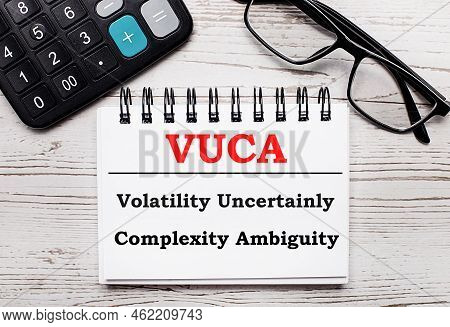 On A Light Wooden Table Calculator, Glasses And A Blank Notepad With The Text Vuca Volatility Uncert