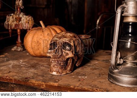 On A Wooden Table Lies A Human Skull, A Pumpkin And A Lamp. Halloween Party
