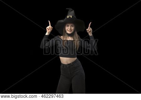 Young Woman In Pointed Hat And Black Clothes Points Fingers Up. Portrait Of Young Witch On Black Bac