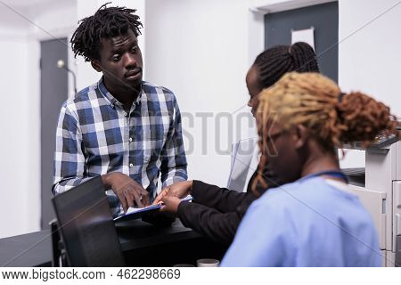 African American Medical Team Talking To Adult At Hospital Reception Desk, Filling In Report Papers 