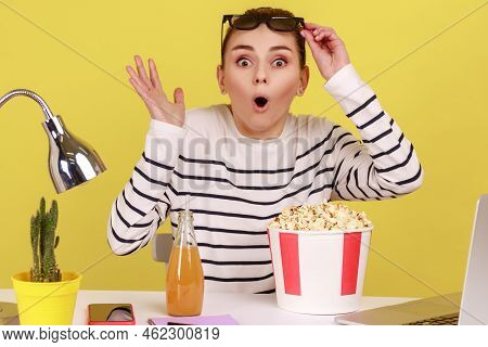 Portrait Of Shocked Impressed Woman Sitting At Workplace With Popcorn And Beverage, Raising Glasses 