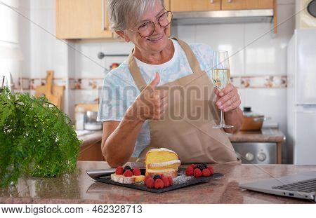 Beautiful Senior Woman In The Home Kitchen After Baking Her Homemade Plumcake, Holding A Glass Of Wi