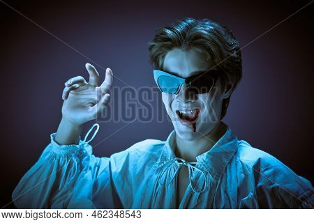 Halloween vampire party. Cool teenage vampire in elegant white shirt and party sunglasses sings and dances with verve. Studio portrait. 