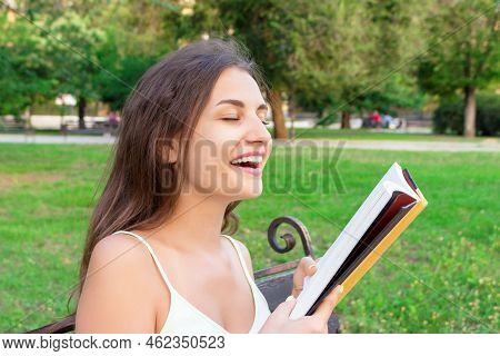 Beautiful Young Brownhair Girl Smells A Fresh Printed Book Sitting On The Bench In The Park.