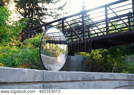 Outdoor View Of Urban Hiking Trail With Convex Safety Mirror, Pedestrian Bridge, And Trees On Bright