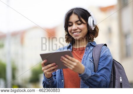 Smiling Middle Eastern Female Student In Wireless Headphones Using Digital Tablet Outdoors